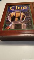 Wooden box with Clue game cover artwork of mansion with characters visible through a magnifying glass.