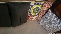 Five decorative pillows arranged side by side on floor, showing various colors and patterns including gray, red and blue/white stripes, and multicolor circle pattern.