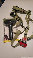 Grooming brush in packaging labeled Miracle Coat large size, adjacent to a tan adjustable dog harness with black and red plastic components on a white surface
