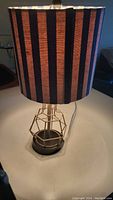Full view of lamp showing striped blue and orange fabric shade and geometric gold-tone metal base on black platform, lamp is turned on showcasing warm light.