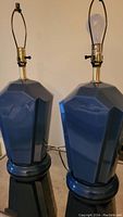Photo showing two solid glass blue table lamps with brass sockets and harps, standing side by side on a flat surface.