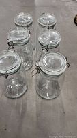 Photo showing 6 large clear glass Mason jars with metal clamp lids on a concrete floor, taken from a slightly elevated angle.