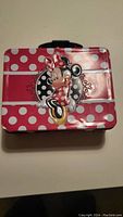 Front view of pink tin lunch box featuring Minnie Mouse with polka dots and decorative details.