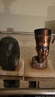 Two vintage Egyptian copper paperweights displayed on a reflective glass surface; a Queen Nefertiti bust on an ivory base and a King Tut pharaoh bust on a similar base.