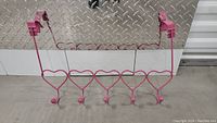 Pink metal over-the-door towel holder with five heart-shaped hooks, lying flat on surface.