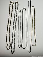 Six assorted silver tone chains laid vertically on a white surface to show variety in design and length.