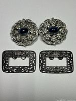 Front view of two oval shoe clips with blue cabochon stones and rhinestones above two rectangular ornate belt buckles.