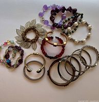 Collection of bracelets and bangles laid out on white background showing various styles and materials in purple, red, gold, and silver tones.