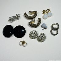 Top view of eight assorted pairs of clip-on earrings displayed on white surface showing variety in size, style, and design.