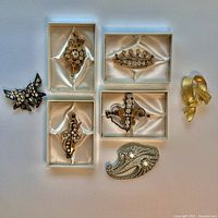 Seven vintage decorative pins and brooches arranged on white background, some in satin-lined boxes with small chains
