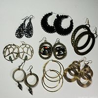 Top view of seven pairs of assorted costume jewelry earrings on white background showing variations in style including black bead hoops, gold-tone geometric and oval designs, and faceted gem accents.