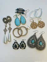 Overview of seven pairs of earrings including turquoise teardrops, pearl drops, gold and silver hoops, black rhinestone drops, and gold textured discs.