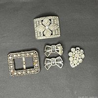 Five assorted metal clips on a black background including rectangular rhinestone shoe clips, bow-shaped clips, and a floral cluster pin.