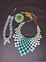 Three costume jewelry necklaces: multi-strand pink and gray beaded necklace, turquoise and white beaded bib necklace, green circular pendant necklace with gold-tone cross and bird motif on chain.