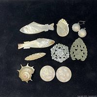 Top-down view showing entire lot of assorted carved pieces on black background. Includes fish shapes, shell shape, carved floral shapes, earrings, round pieces with swirl patterns, and leaf-shaped piece.