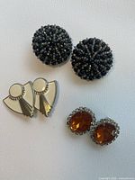 Close-up photo of three pairs of earrings: beaded black dome-shaped with gold accents, cream and gold geometric fan-shaped, and amber-colored oval rhinestone surrounded by clear rhinestones.