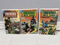 Three 'Unknown Soldier' comic books from DC Comics shown side by side, each bagged and boarded. The first comic is issue #208 dated October 10, 1977, the second is issue #215 dated May 10, 1978, and the third is issue #238 dated April 10, 1980. Covers depict military combat scenes with soldiers, helicopters, and weaponry.