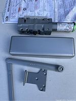 Photo of commercial door closer parts including main body, cover, arms, mounting plate and instruction paper on a white surface