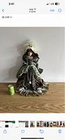 Front view of Victorian porcelain doll wearing black polka dot and green layered satin dress with lace and ruffles, feathered hat, curly brown hair; includes soda can for size reference