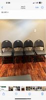 Four black metal folding chairs with dark gray padded seats and backrests lined up against a wall on hardwood floor.