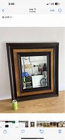 Photo of rectangular mirror with wooden frame resting on a wooden floor against a white wall, showing reflection of a room with bicycle and blue bag.