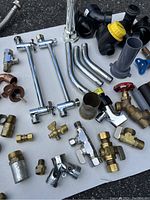 View of plumbing materials including brass valves, chrome pipe fittings, copper pipes, and plastic valves arranged on a surface.