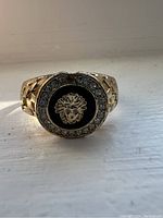 Front view of gold tone ring showing black Medusa emblem center surrounded by rhinestones.