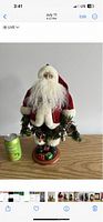Front view of a Santa Claus figurine standing on a wooden base with green artificial pine decoration. A soda can is placed next to the figure for size comparison.