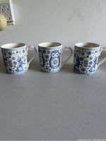 Three white ceramic cups with detailed blue floral and geometric patterns on their exterior, each with a handle.