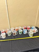 Photo showing the full set of 16 Fireking mugs arranged in a row against a wall on a black cloth surface with a yellow measuring tape in front for scale.