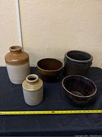 Five ceramic pottery pieces including jugs, bowls, and an urn, arranged on a black surface with measuring tape.