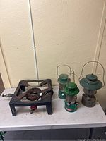 Gas stove and three Coleman gas lanterns placed on a white table against a beige wall, showing overall lot contents.