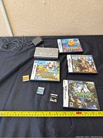 Photo showing silver Nintendo DS console with charger and five Nintendo DS game cartridges and cases arranged on black surface. Measuring tape included for scale.
