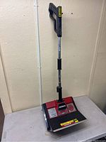 Full view of red and black NOMA Turbo Shovel electric snow shovel standing on table against a wall, showing handle and scoop.