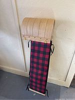 Full view of the old wooden snow sled showing curved wood slats on front and red and black checkered padded seat.
