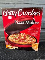 Front view of the boxed pizza maker showing the Betty Crocker brand, product name, and an image of a cooked pizza with some cheese beside it.