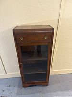 Front view of wooden china cabinet with single drawer and glass door showing interior shelves.