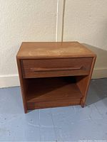 Front view of teak nightstand showing drawer and open shelf, with surface wear on top.