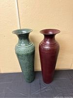 Photo showing two tall metal vases side by side, green and reddish-brown with flared necks and embossed decorative bands.
