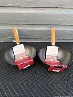 Two new 8-inch Palm professional quality woks shown side by side with packaging labels attached, placed against a grey wall on black quilted surface.