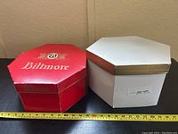 Two octagonal hat boxes, one red Biltmore branded, the other white with gold trim, measured next to a tape measure showing size approximately 12 inches across.