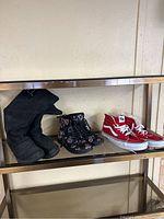 Three pairs of women's shoes on a glass shelf: black suede boots, black floral lace-up boots, and red Vans sneakers.