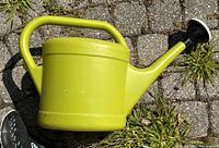 Lime green plastic watering can showing side profile on pavement with grass.