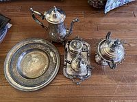 Full silver tea set on wooden floor, showing tea pot, warming stand, creamer, trays, and sugar bowl with bird finial lid.