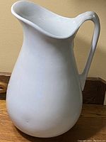 White ceramic pitcher with large curved body, wide spout, and detailed handle on wooden surface.