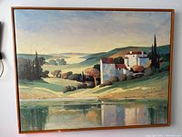 Full view of framed oil painting showing scenic landscape with houses by a lake.