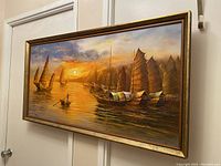 Framed oil painting showing a sunset scene with several sailboats and warm colors, hung on a white wall.