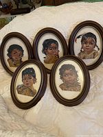 Five oval framed petit point needlepoint artworks showing Native American children arranged on a quilted white surface.