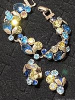 Close-up of bracelet and earrings showing blue and champagne rhinestones and silver-tone leaf accents on black fabric background.