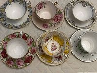 Top view showing all six cups with their matching saucers arranged on a quilted surface, different floral and star patterns visible with gold rims.